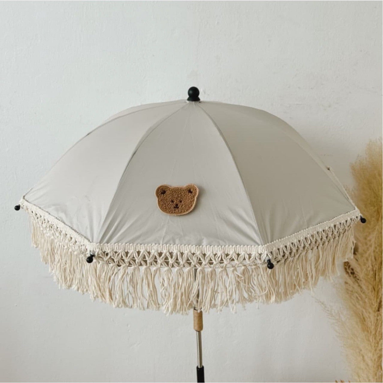 Beige umbrella with teddy bear emblem and fringe details on a light gray background