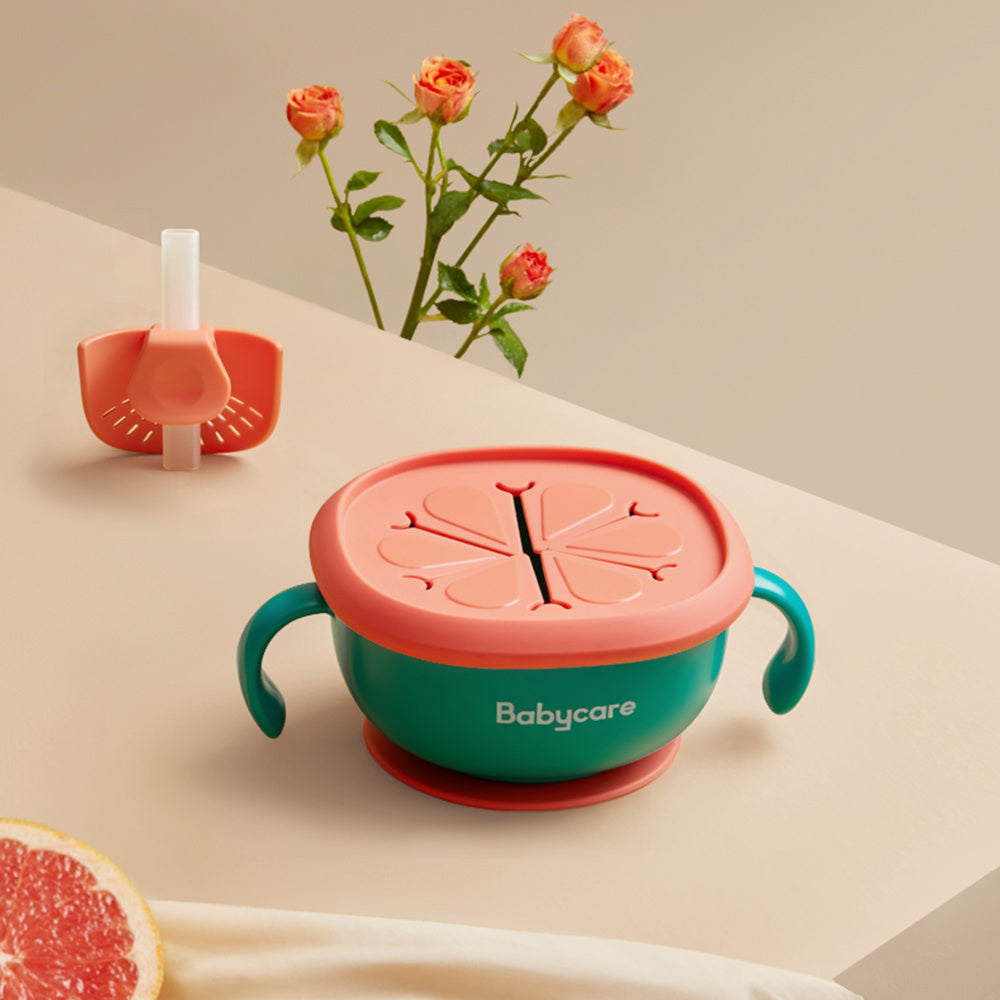 Colorful baby bowl with pink lid and green handles on a beige surface with flowers and fruit.
