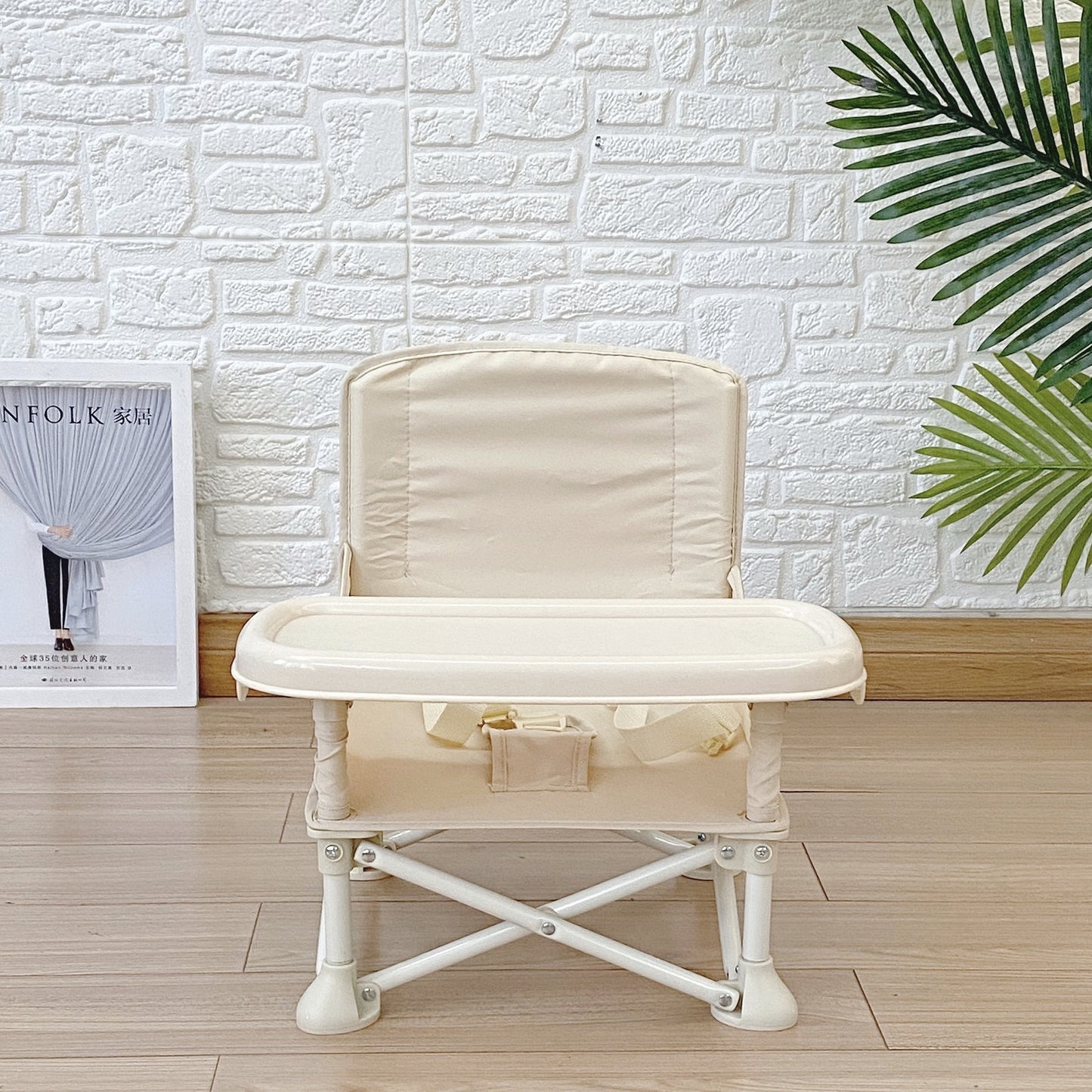 Beige baby high chair against a white brick wall with a plant and magazine in the background.