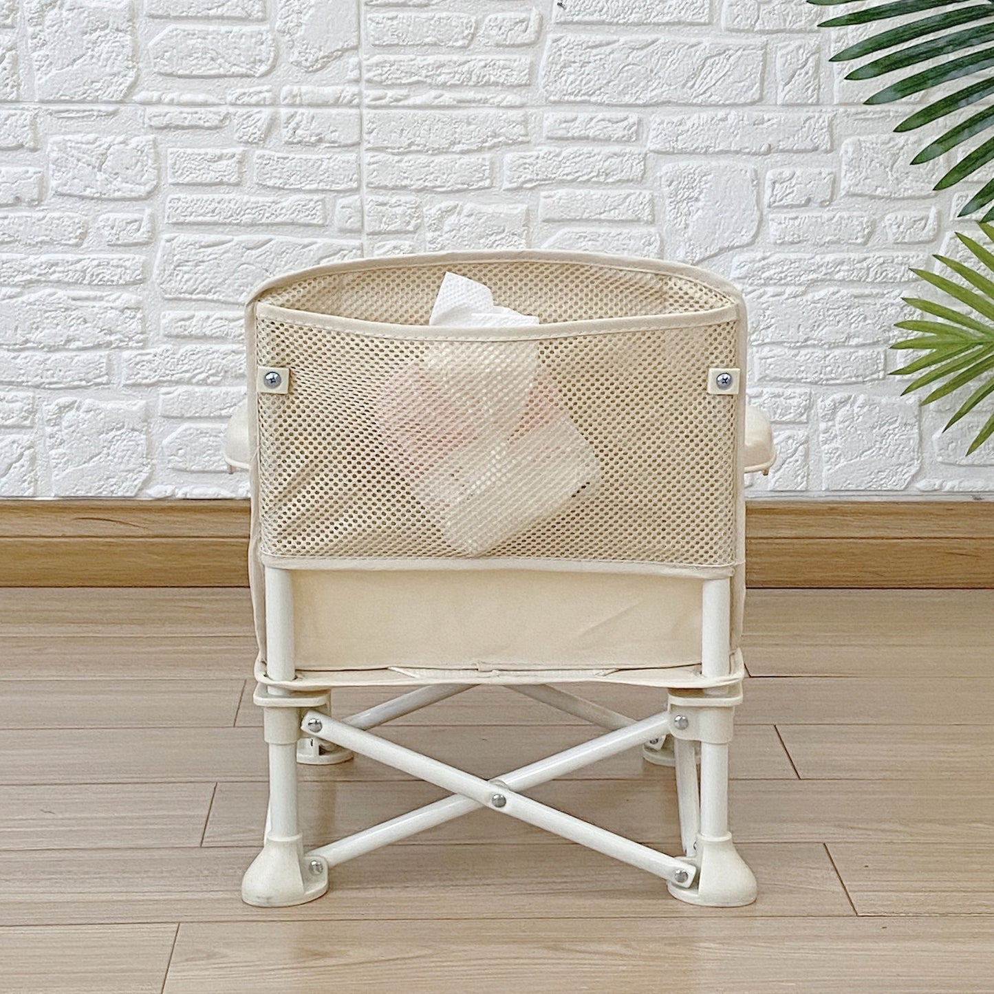 Beige mesh baby crib with white base against a light brick wall.