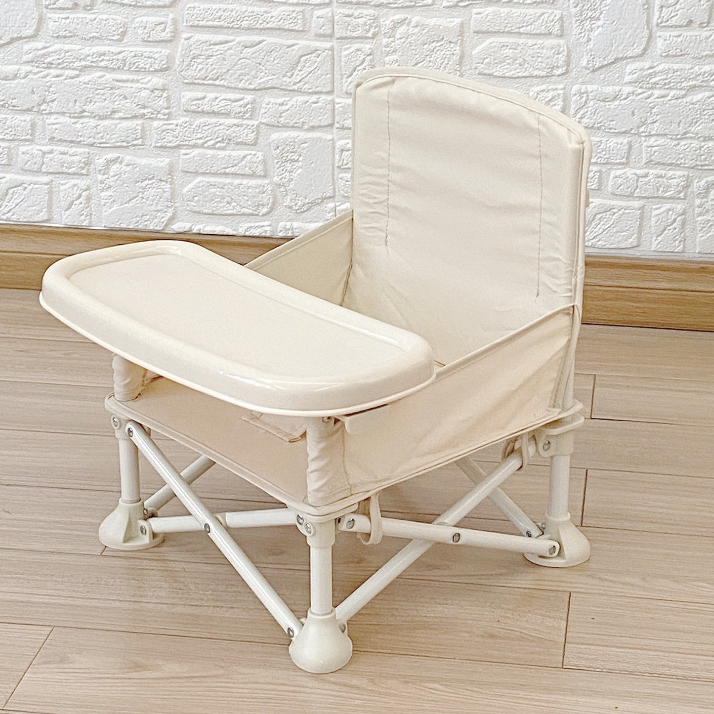 Beige baby high chair with tray on a wooden floor against a white brick wall.
