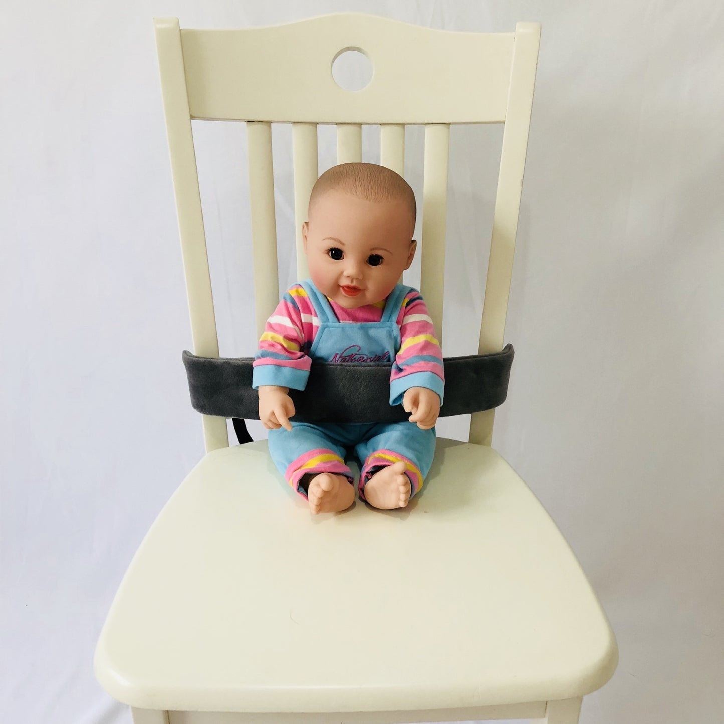 Baby doll in a colorful outfit sitting on a chair with a seat belt around it.