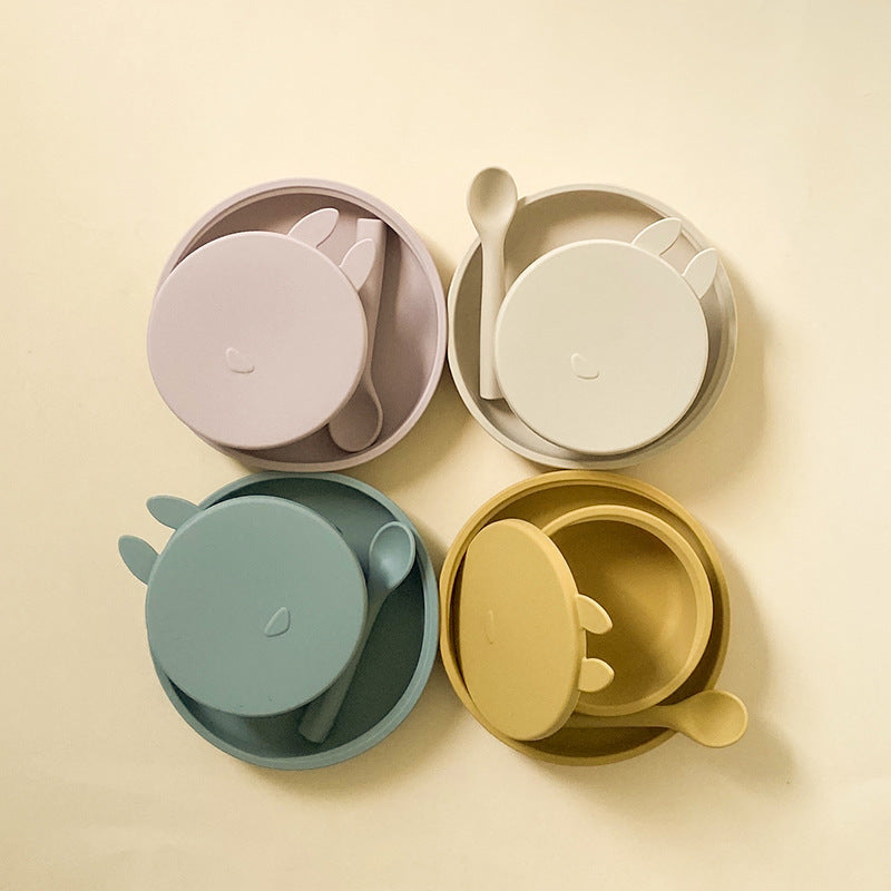 Set of four children's bowls with lids and spoons in pastel colors on a beige background