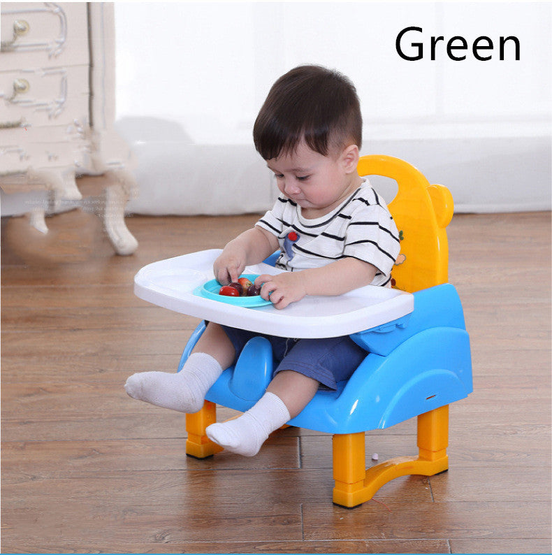 Child sitting in a blue and yellow booster seat with a plate of food on a wooden floor.