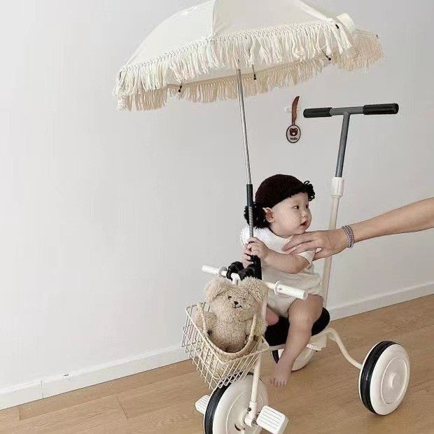 Child sitting on a tricycle with a teddy bear in a basket and an adult's hand holding the handle, against a plain white wall.