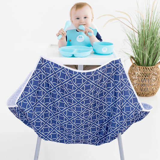 Baby in a high chair with blue bib and utensils, wearing a blue bib with geometric pattern.