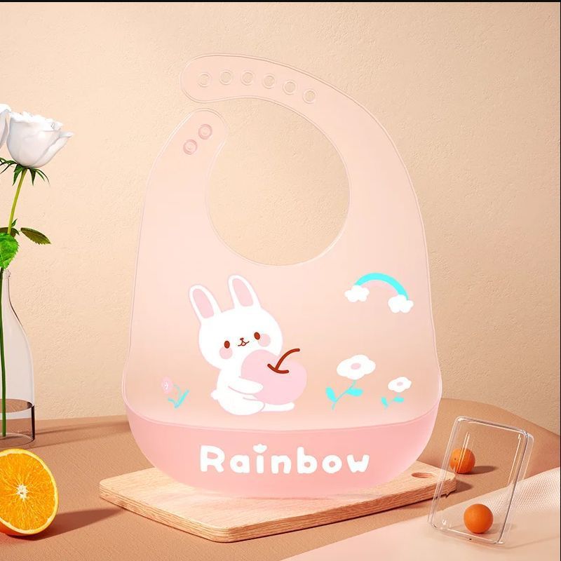 Pink children's bib with cartoon character and 'Rainbow' text on a beige surface.