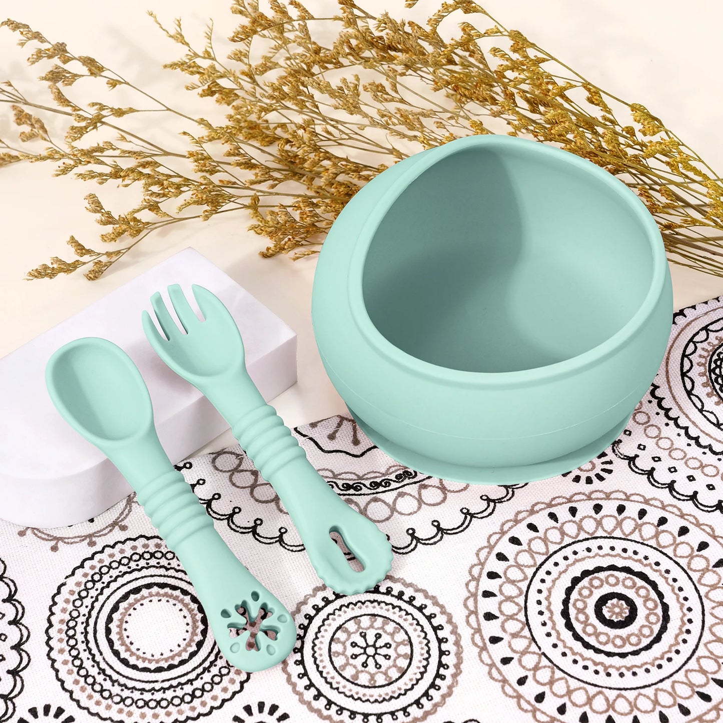 Light blue silicone bowl and utensils on a decorative mat with gold flowers.