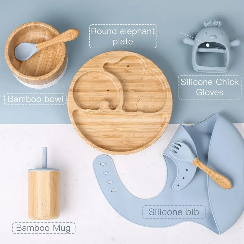Collection of bamboo and silicone baby products including a bowl, plate, mug, bib, and gloves on a light blue background.