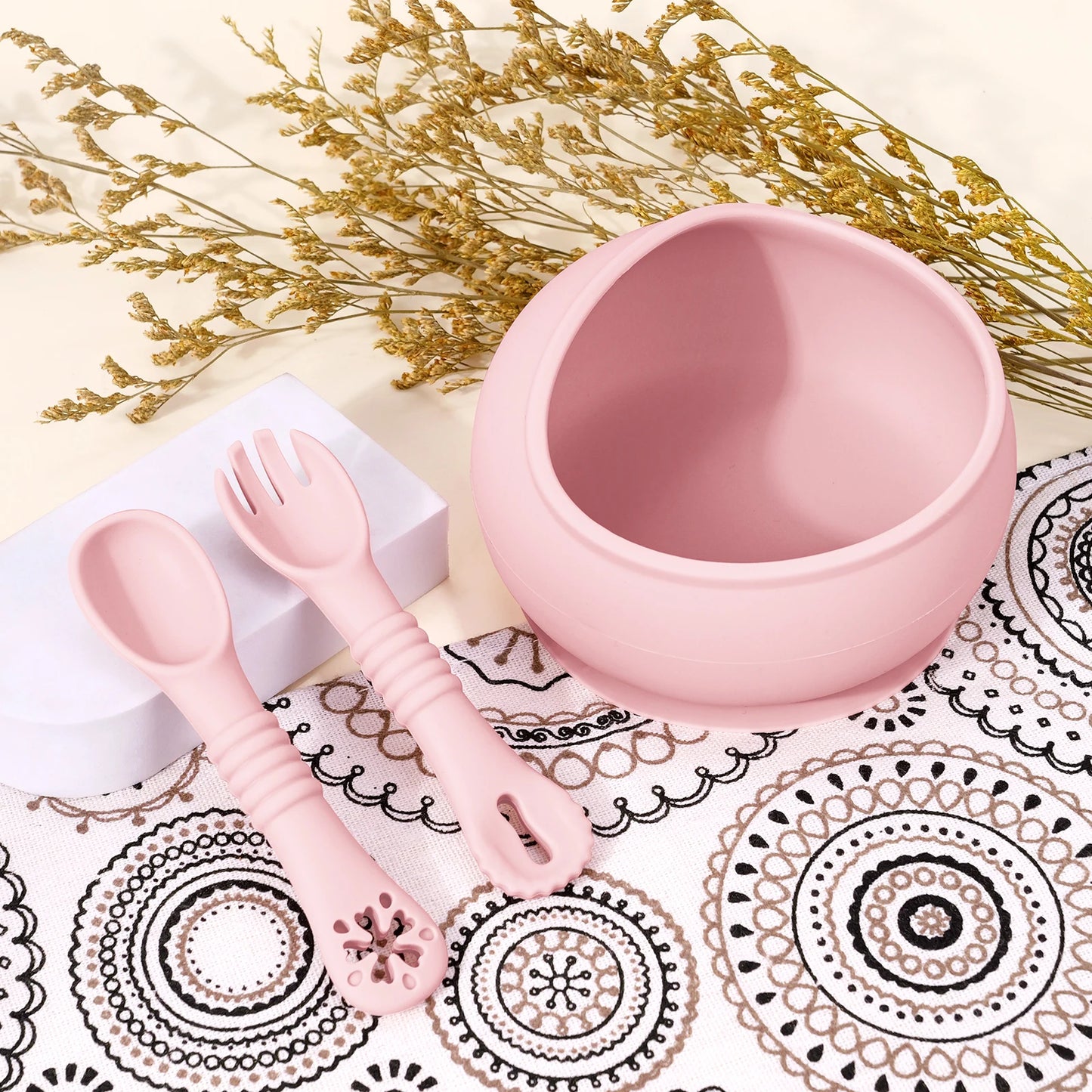 Pink children's bowl, spoon, and fork set on a decorative tablecloth with gold flowers.