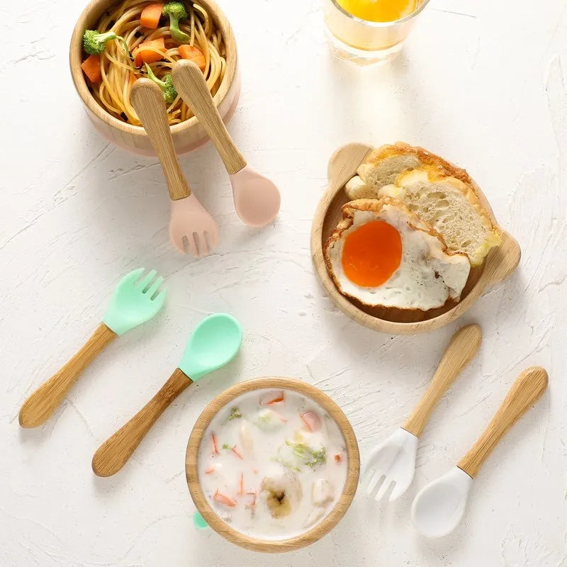Children's wooden and silicone utensils with a bowl of soup, plate of bread and egg, and a small bowl of noodles on a white surface.