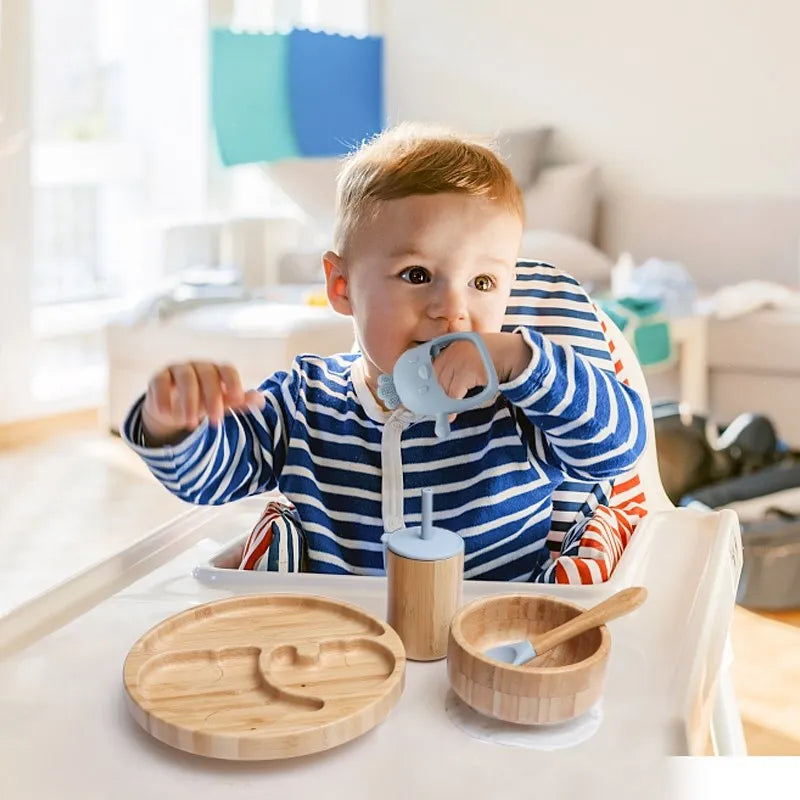 Child in a high chair with wooden餐具 and a teething ring, sitting in a bright room.