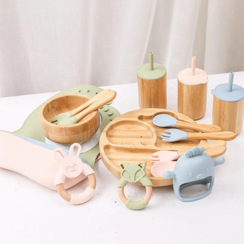 Collection of wooden and silicone baby utensils and toys on a white surface with a light gray curtain background.