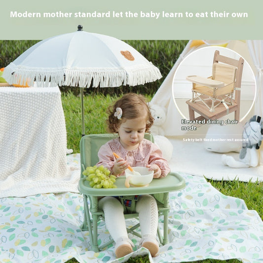Child sitting in a portable high chair outdoors with an umbrella and picnic blanket.