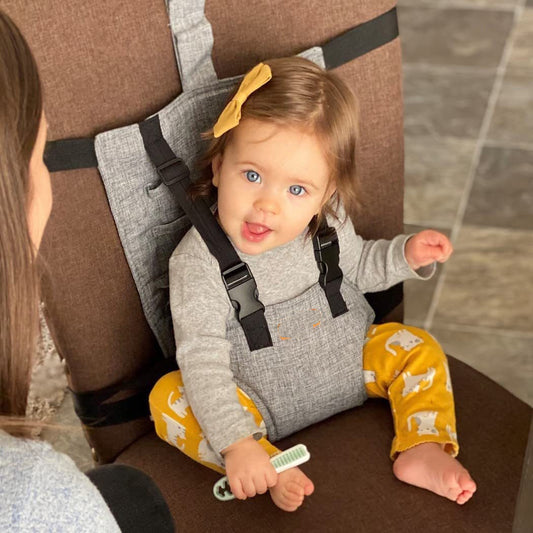 Child in a gray baby carrier with yellow pants and a bow, sitting on a brown chair.