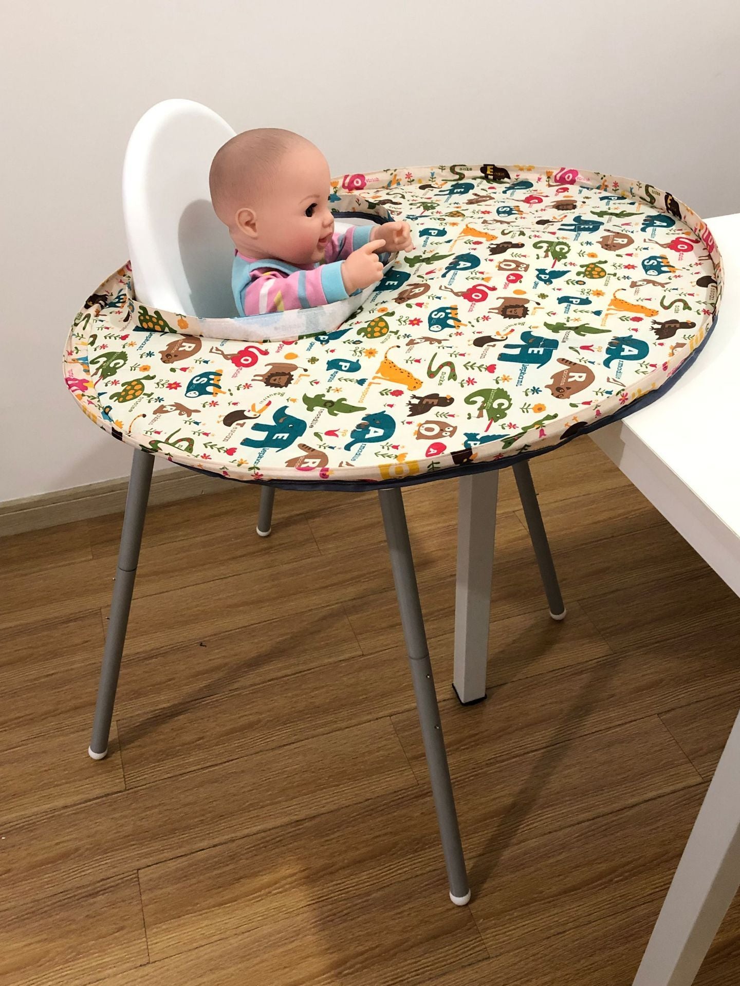 Round table with colorful animal print cover and baby in high chair on a wooden floor.