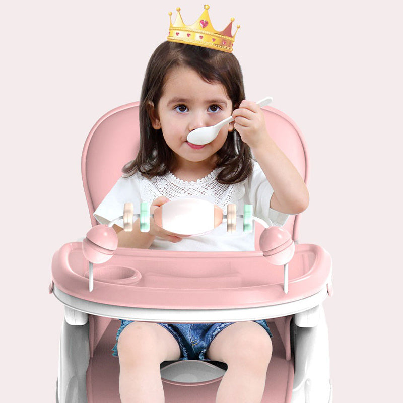Child sitting in a pink high chair with a crown on, holding a spoon.