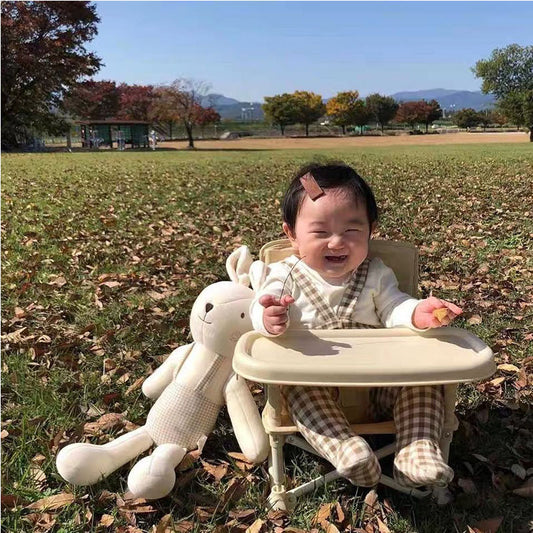 Baby sitting in a high chair with a stuffed toy outdoors on a grassy field.