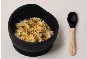 Black bowl with rice and vegetables, wooden spoon, and black spoon on a light gray background