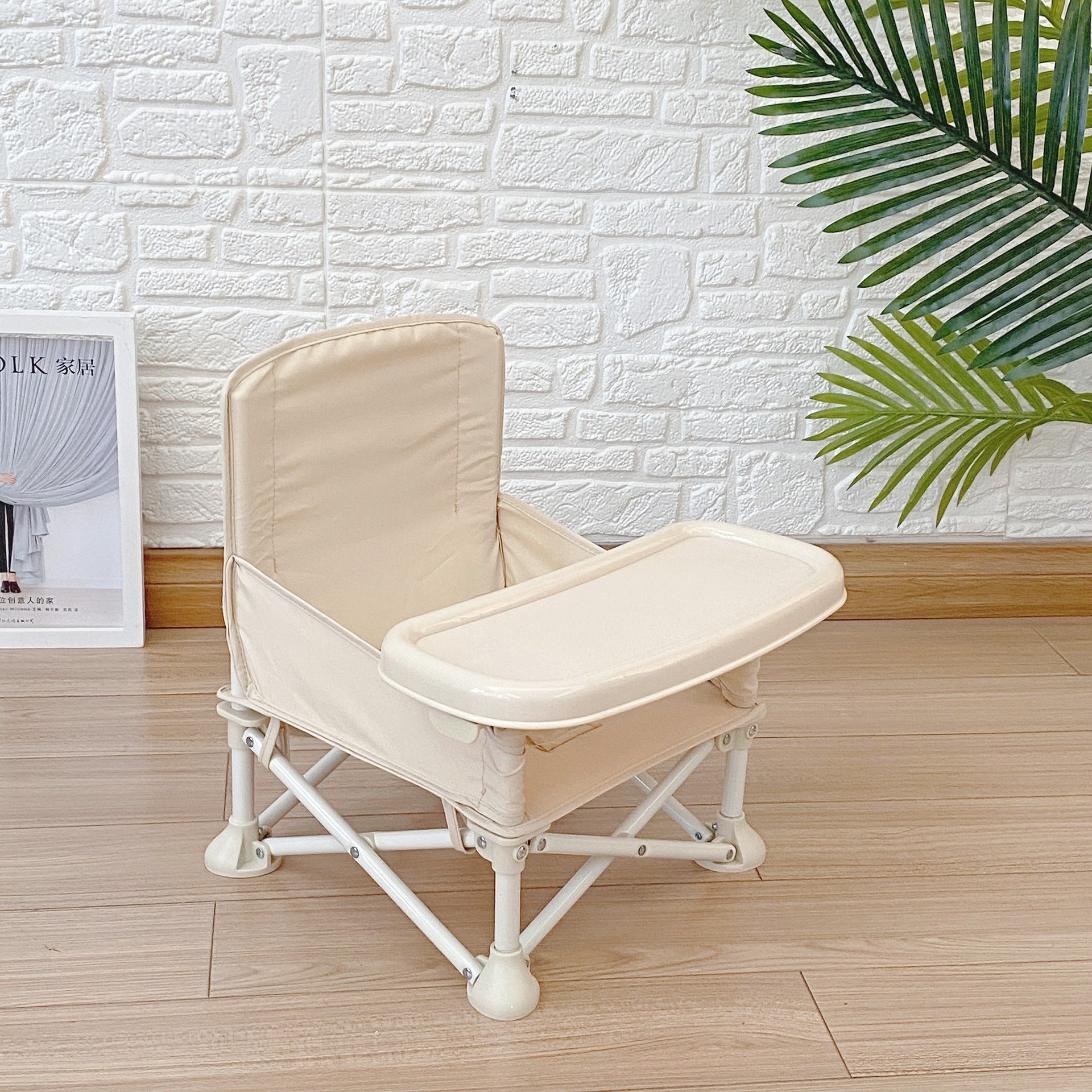 Beige baby high chair on a wooden floor with a white brick wall and green leaves in the background