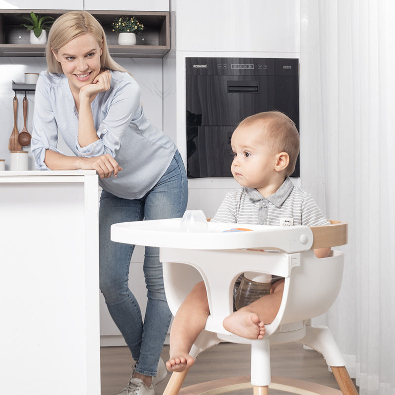 Woman standing next to a child in a high chair in a kitchen.
