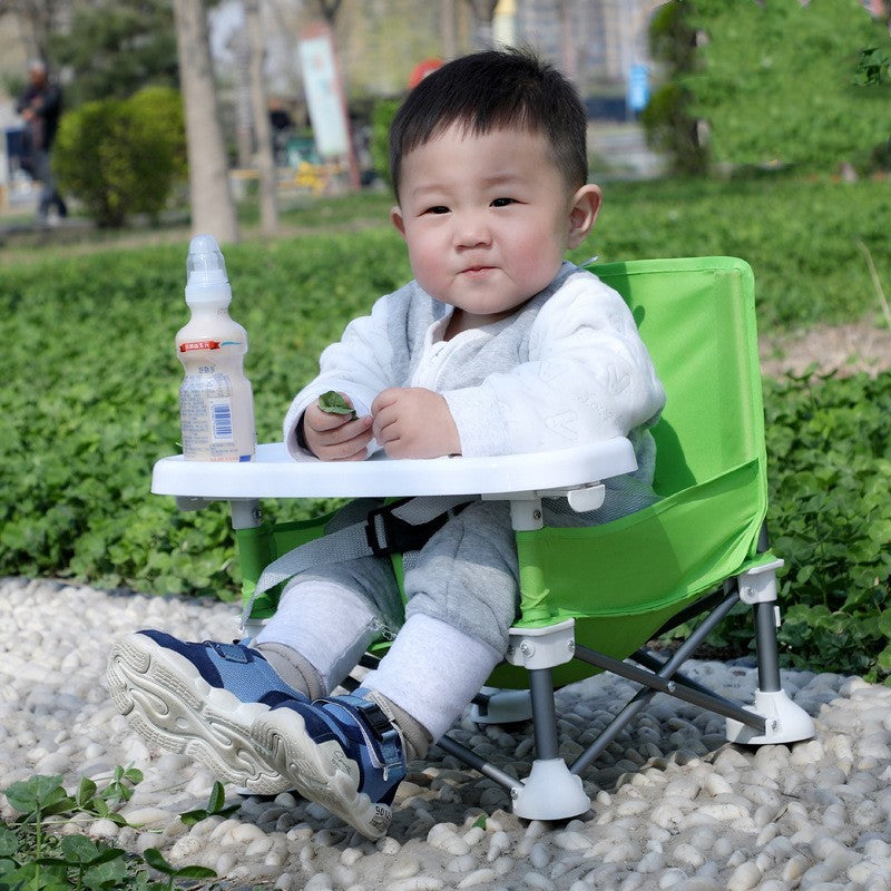 Child sitting in a green and white folding chair outdoors with a bottle on the tray.