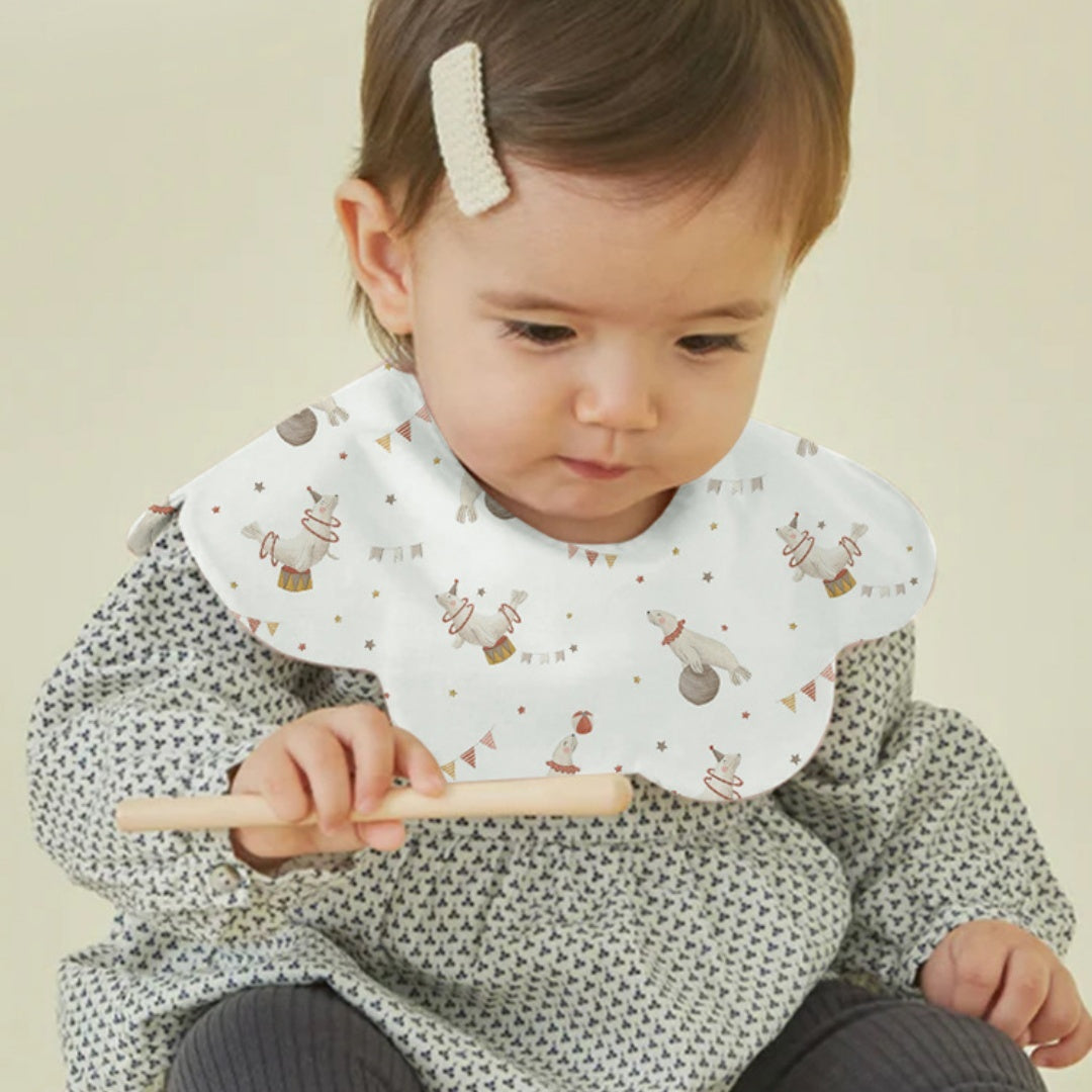 Baby wearing a patterned bib and holding a wooden spoon against a plain background