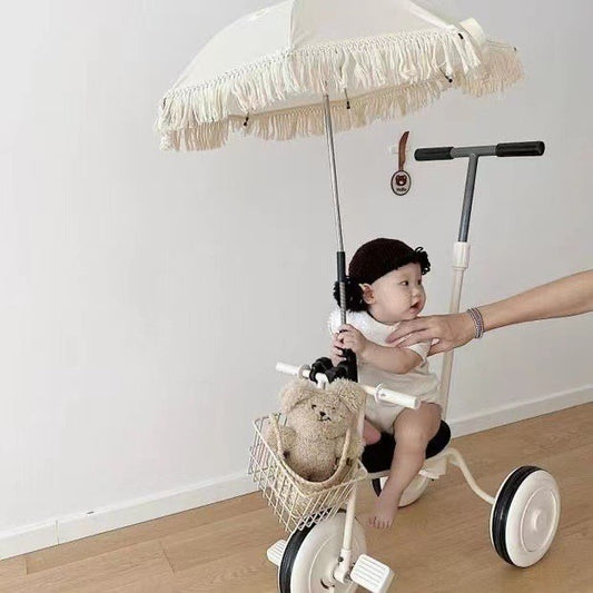 Child sitting on a tricycle with a teddy bear in a basket and an adult's hand holding the handle, against a plain white wall.
