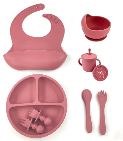 Pink silicone baby feeding set including bib, plate, cup, and utensils on a white background