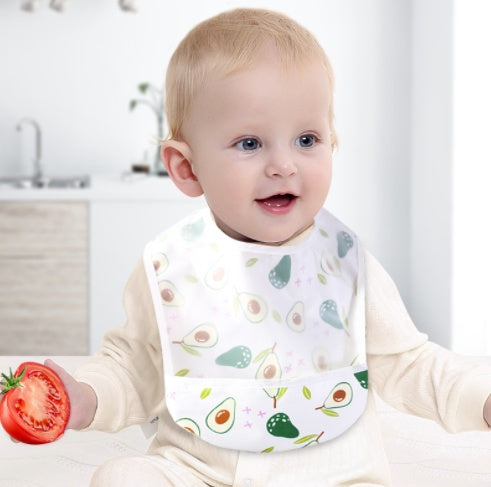 Baby wearing a bib with avocado pattern holding a tomato in a kitchen setting
