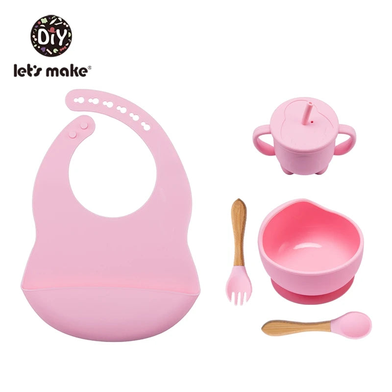 Pink baby utensil set with bib, cup, bowl, fork, and spoon on a white background.
