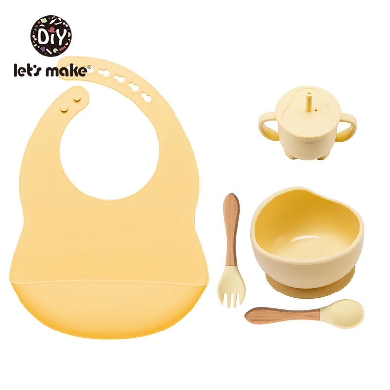 Yellow baby bib, cup, bowl, spoon, and fork set with 'let's make' branding on a white background.