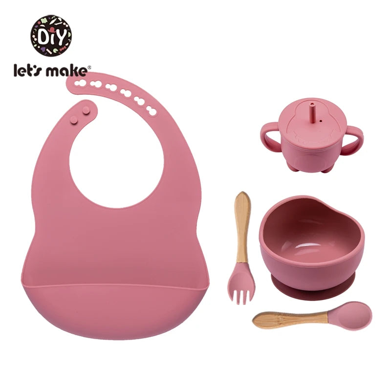 Pink baby feeding set with bib, cup, bowl, spoon, and fork on a white background.