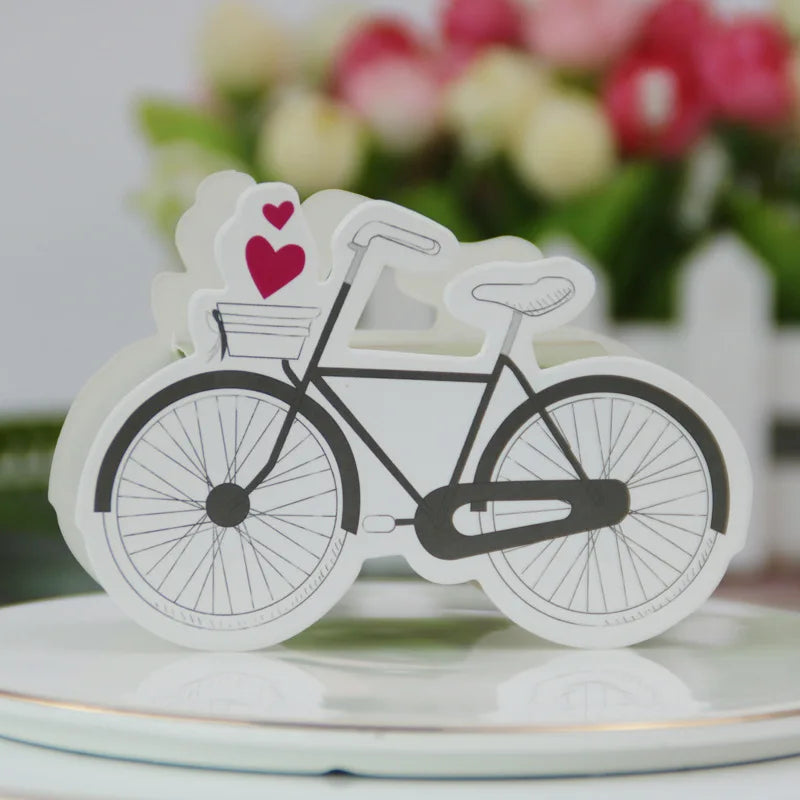 White bicycle-shaped card with red hearts on a white surface with blurred flowers in the background