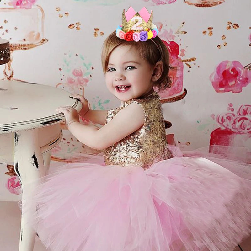 Child in a gold sequin top and pink tutu with a colorful number 2 crown, sitting in front of a floral backdrop.