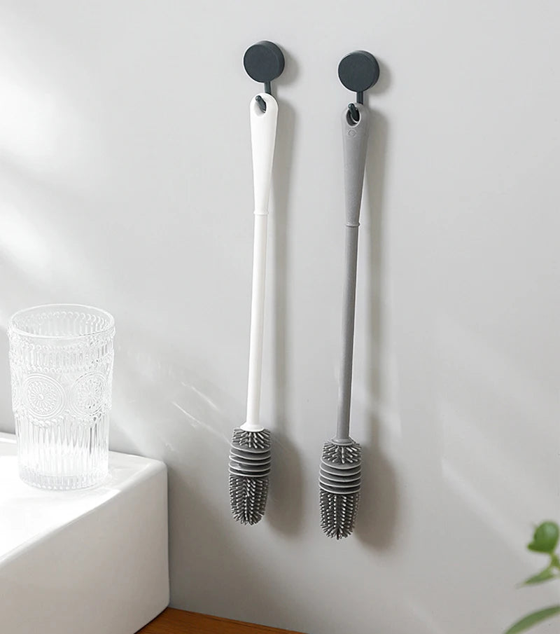 Two bottle brushes with black bristle heads hanging on a white wall next to a glass.