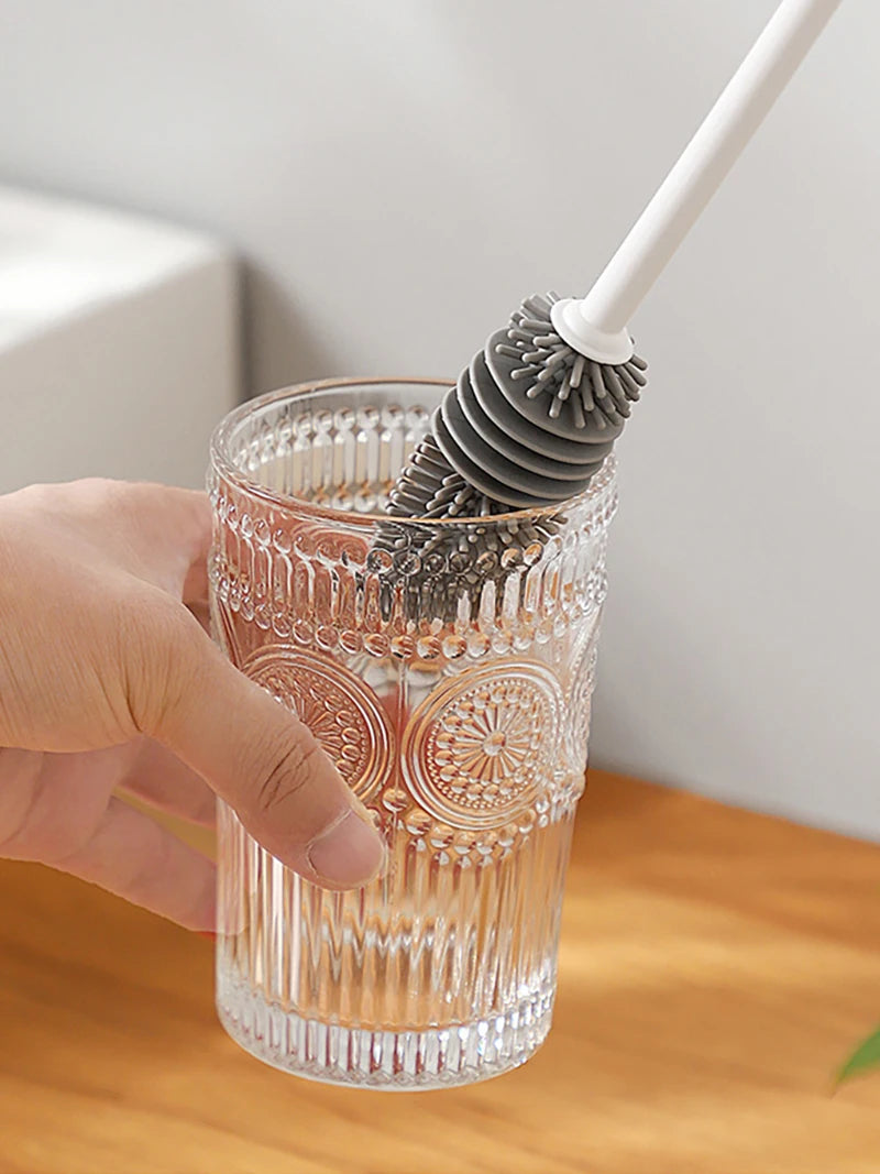 Hand using a silicone bottle brush to clean a glass on a wooden surface.