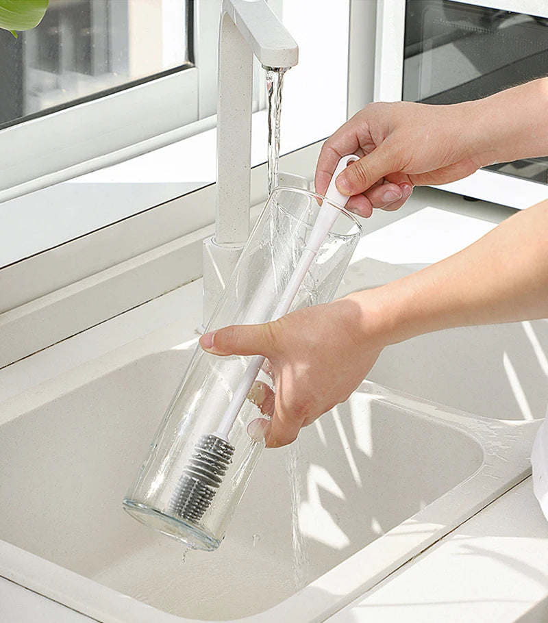 Person cleaning a glass bottle with a brush under running water in a sink.
