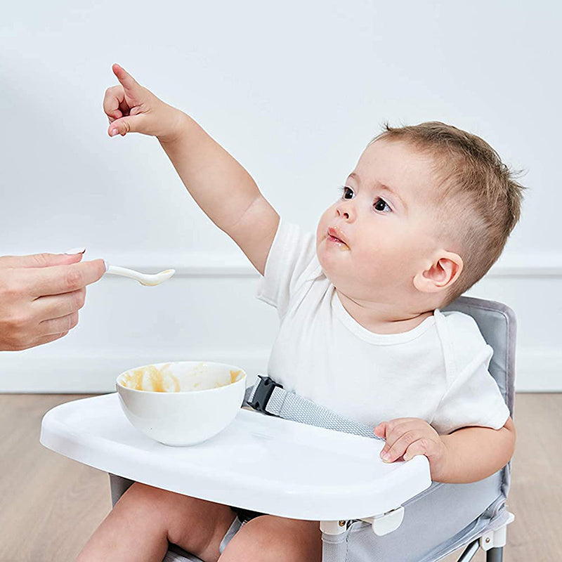 Baby in a high chair being fed with a bowl and spoon against a plain background