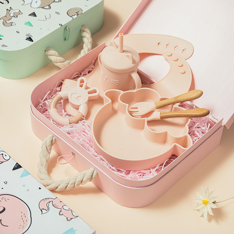Pink baby utensil set with bib, cup, and toys in a box on a light pink background.